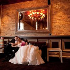 Image of bride and groom posing for pictures with photographer on balcony lounge couches in front of historic exposed brick walls at The Allure Wedding Venue and Event Center